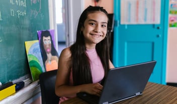 Student working on laptop in classroom smiles at the camera