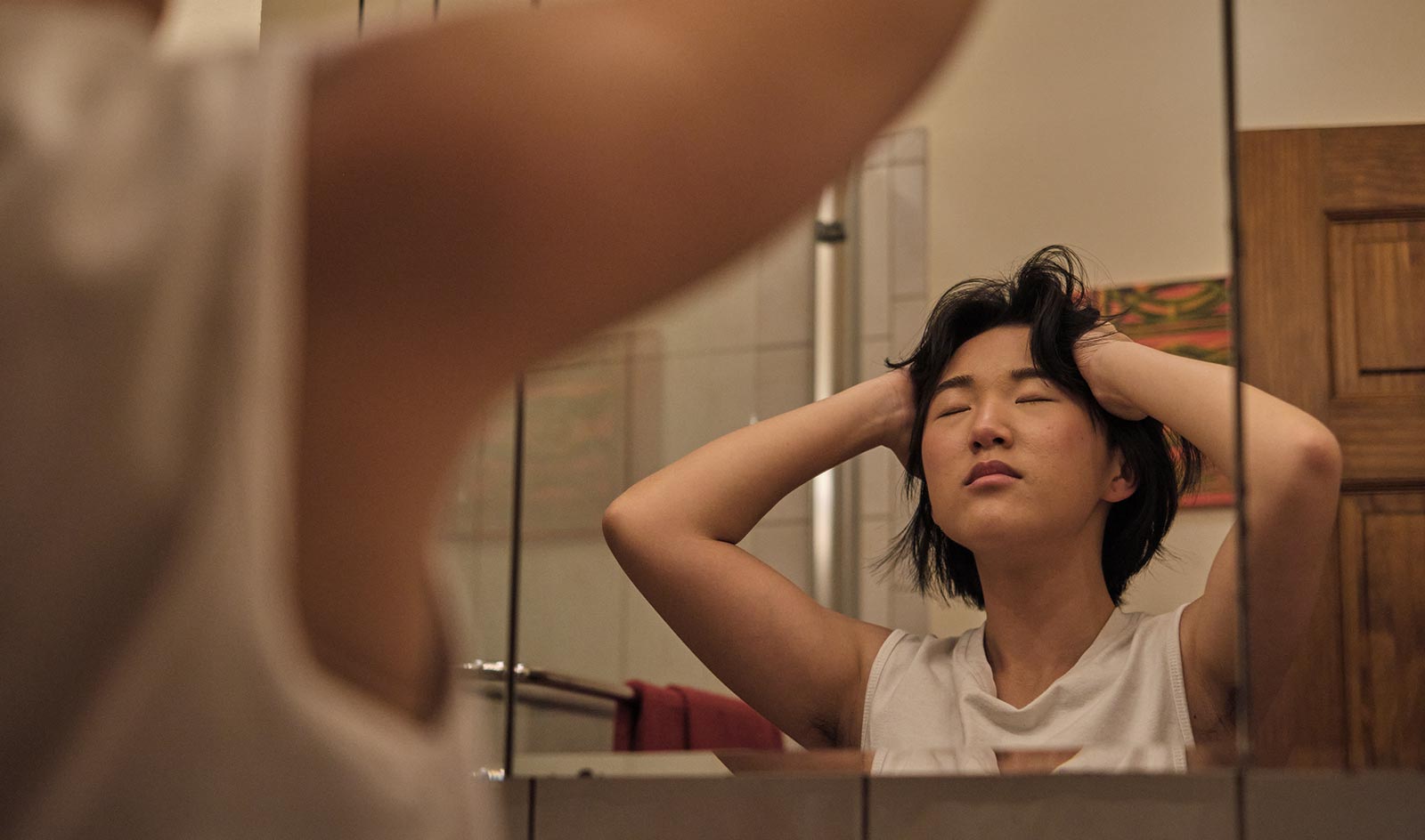 Non-binary person massages head and takes a deep breath facing bathroom mirror