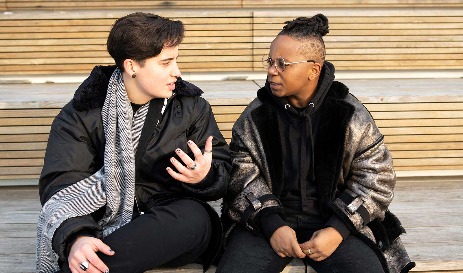 Two trans friends sit on bench and talk to one another