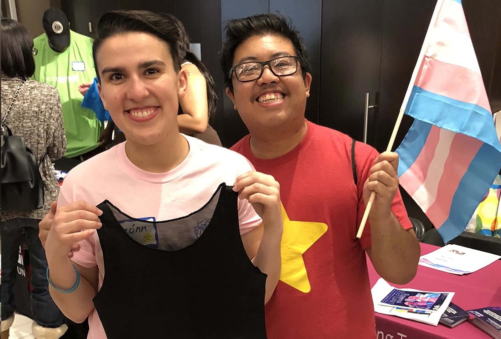 Two smiling trans folks hold up a chest binder